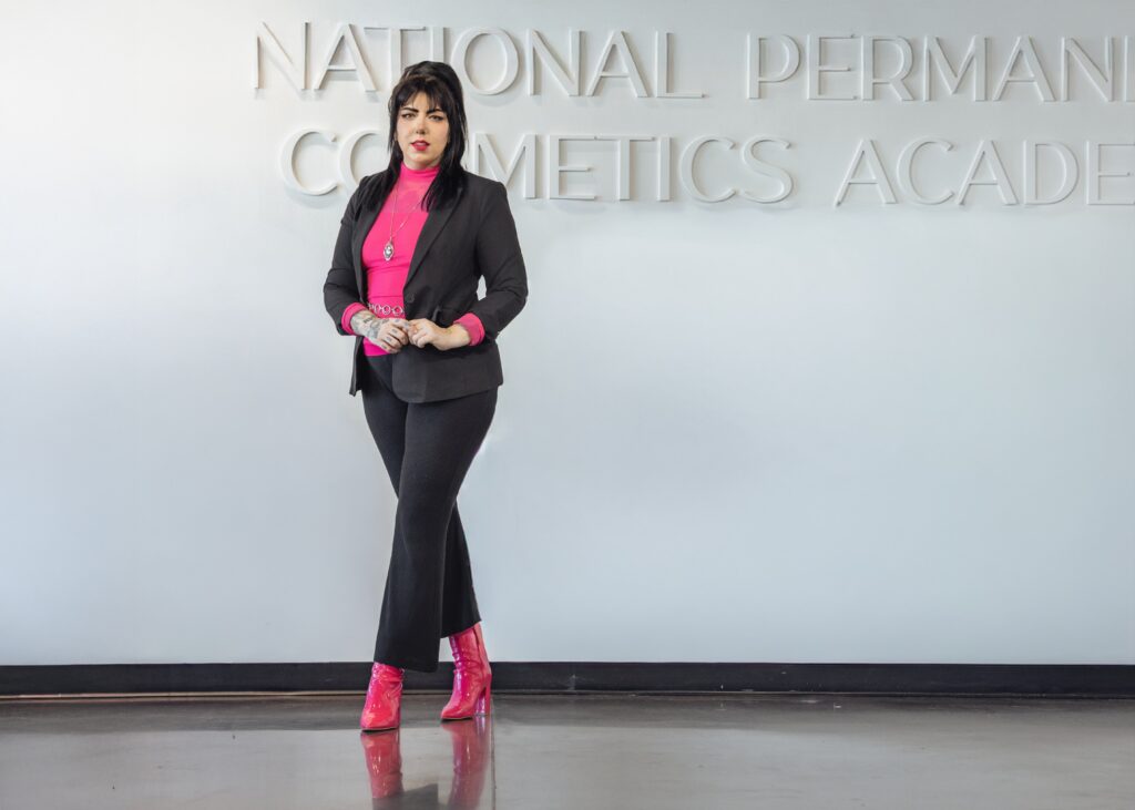 Salt Lake City PMU artist standing in modern studio wearing black suit and pink top with red boots, confident professional branding portrait inside permanent makeup clinic, clean minimal aesthetic, Moonlit PMU Salt Lake City Utah