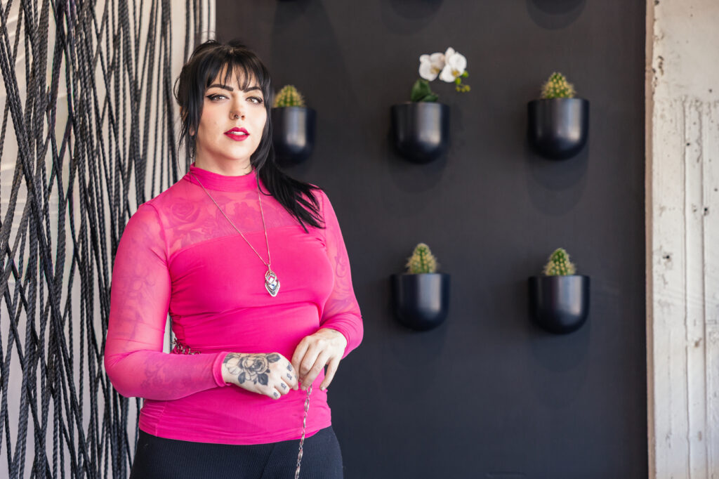 Salt Lake City PMU artist Alison Daigle standing in a modern studio wearing a bright pink top, showcasing confident professional branding portrait with tattoos, styled hair and makeup, natural lighting, Moonlit PMU environment, Salt Lake City, Utah