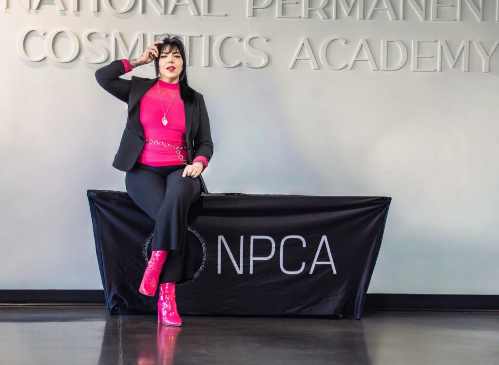 Salt Lake City Utah PMU artist Alison Daigle posing in front of National Permanent Cosmetics Academy sign, wearing bold pink top and boots with black blazer, confident branding portrait for permanent makeup training, cosmetic tattoo artist identity, modern studio environment, high contrast clean lighting, luxury PMU brand presence